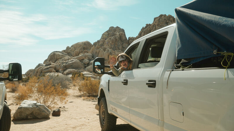 A white pickup truck is parked in a desert landscape with rocky hills in the background. A person is visible in the driver's seat, waving. The truck has a dark-colored covering over the bed. The sky is light blue with some clouds, and the overall scene suggests an outdoor adventure or camping setting.